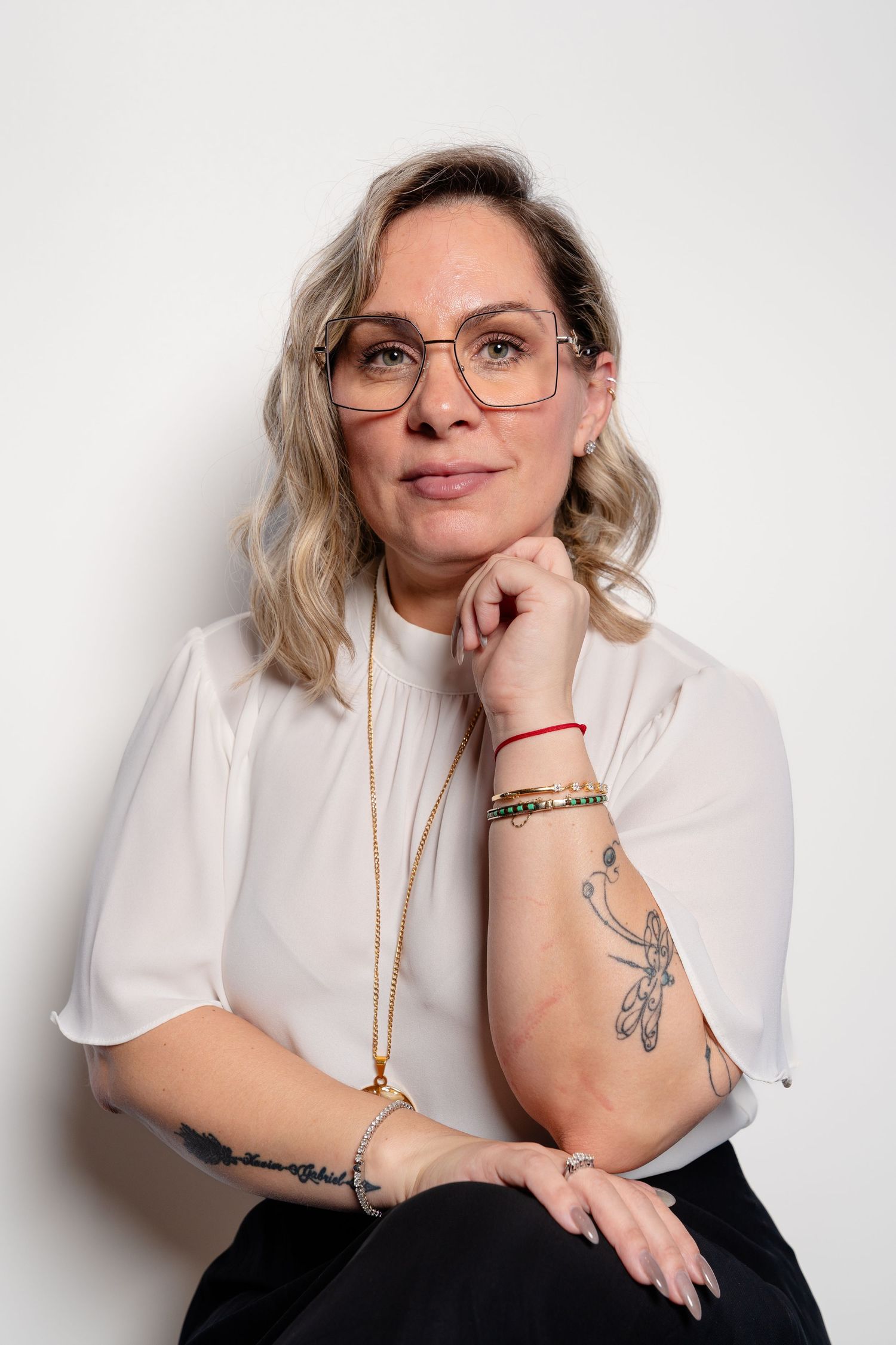 Blonde woman with glasses and tattoos wearing white blouse and gold necklace posing thoughtfully.
