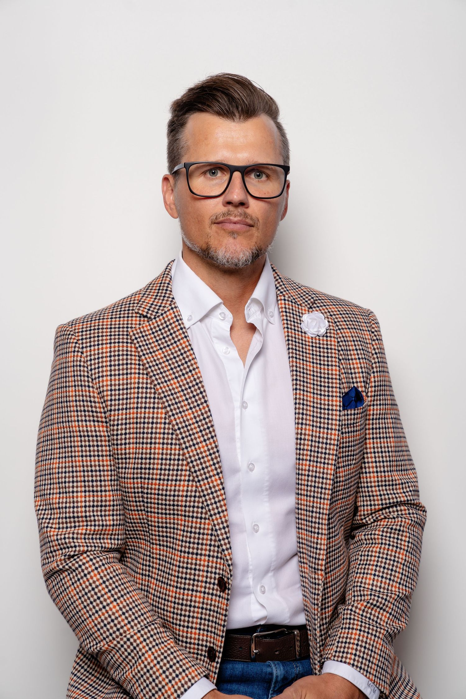 Man in plaid blazer, white shirt, glasses, and blue pocket square poses confidently against white background.