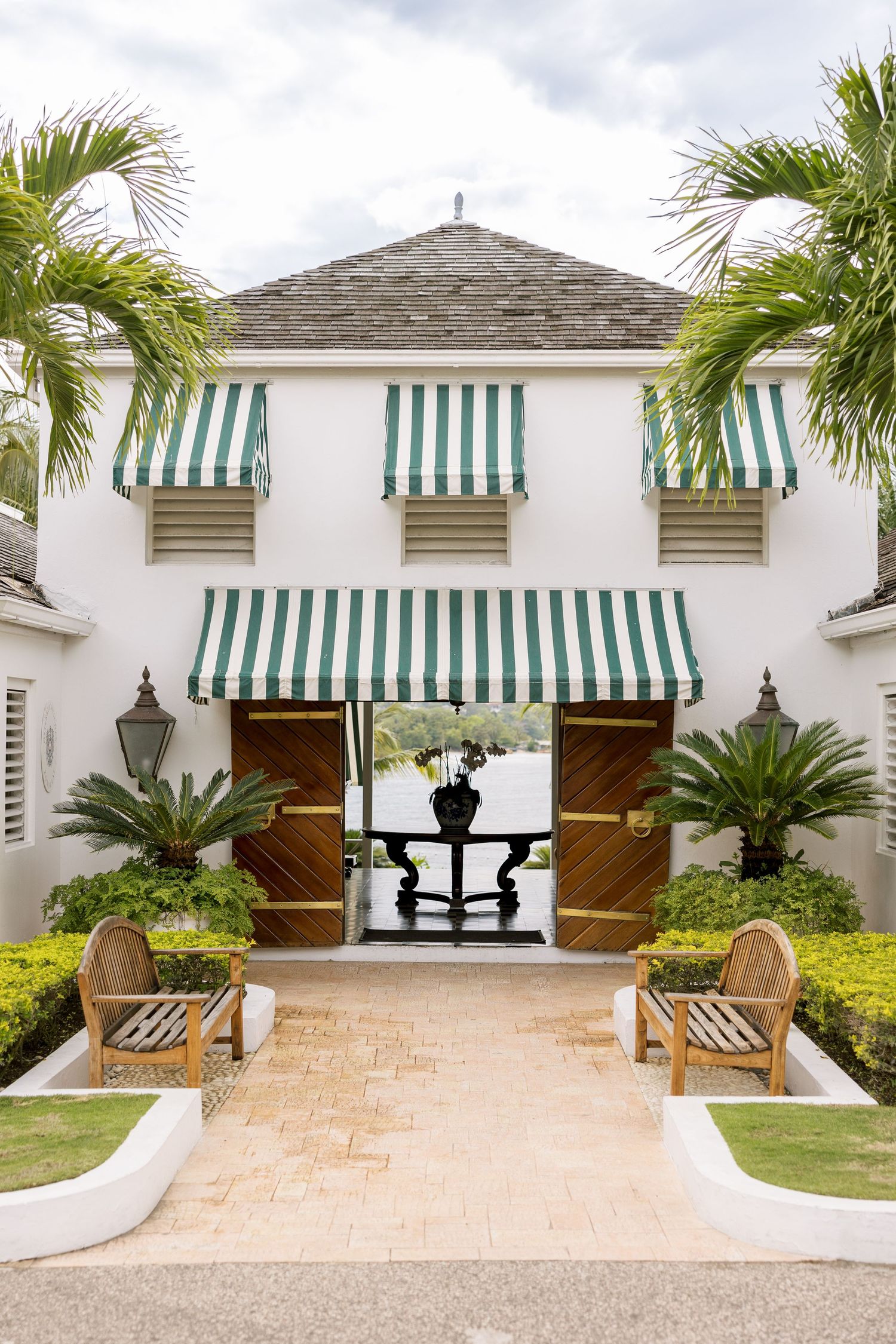 Elegant tropical villa entrance with green striped awnings, wooden doors, palm trees, and manicured hedges on a stone pathway.