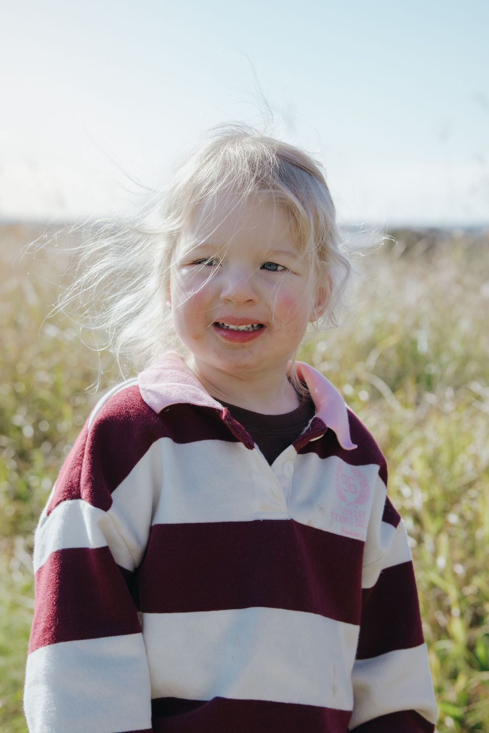 Smiling blonde toddler girl in striped maroon and white shirt standing outdoors in a sunny field with wind-blown hair.