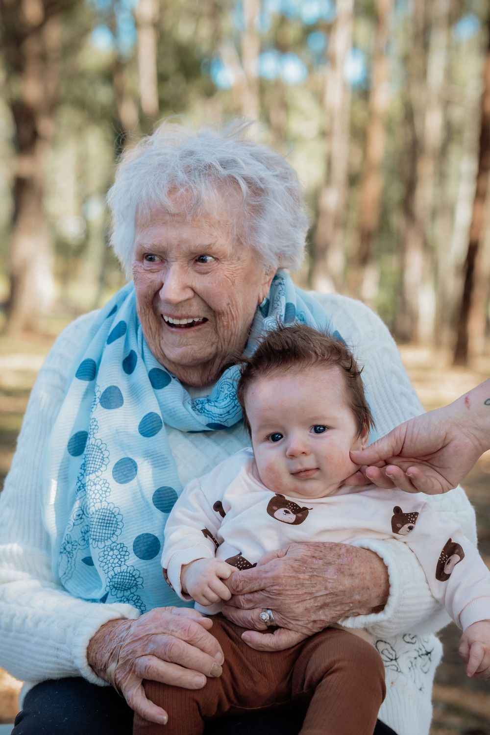 Elderly great-grandmother laughing while holding an adorable baby in bear print onesie outdoors in a forest setting.