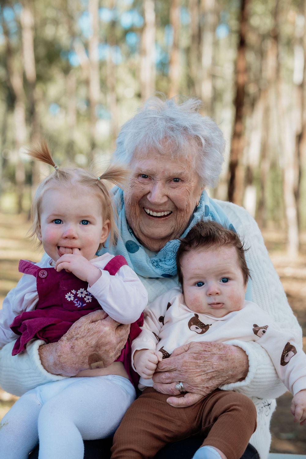 Elderly grandmother smiling warmly while holding two young toddler grandchildren outdoors in a forest setting.