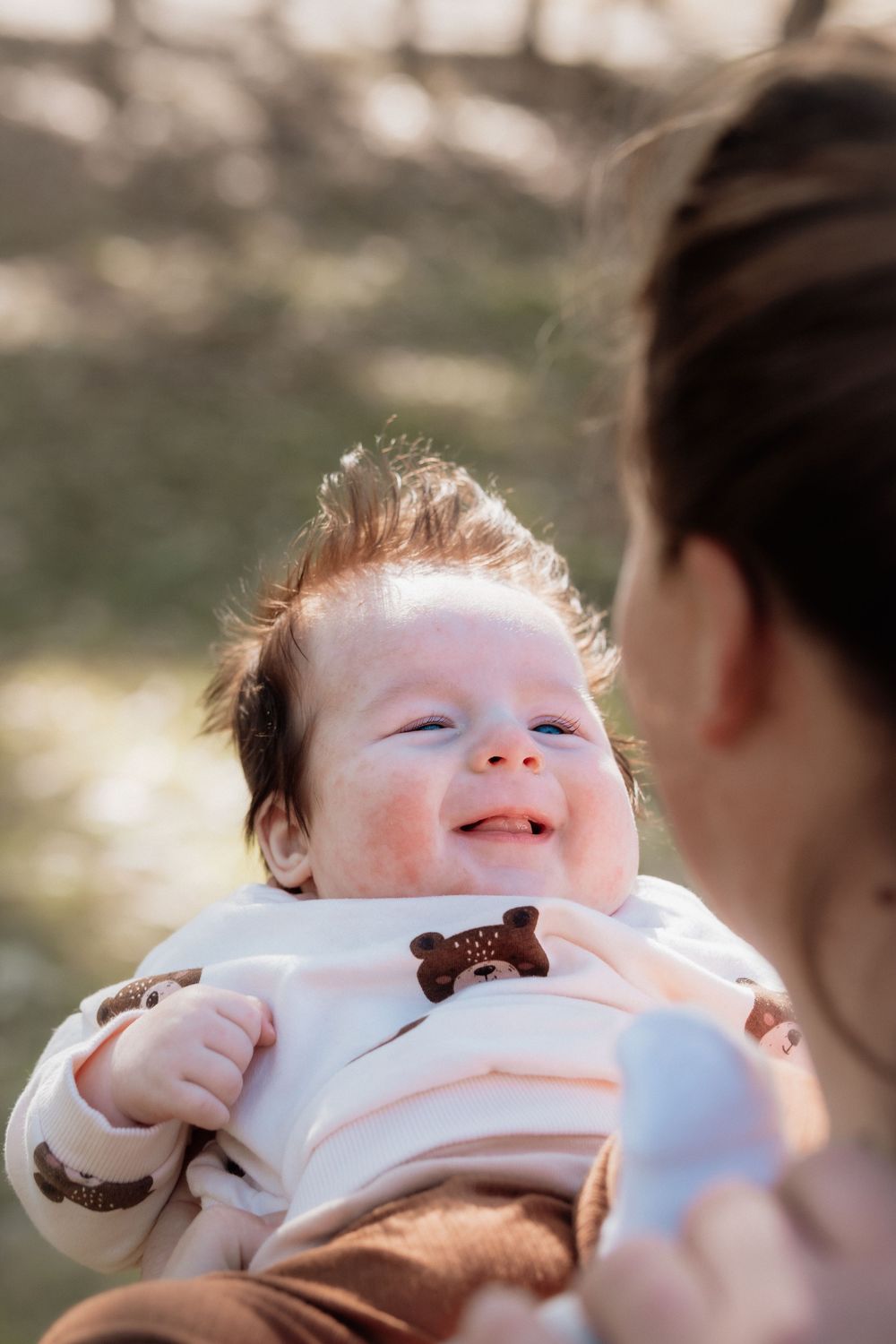 Happy smiling baby with rosy cheeks held by mother outdoors in warm sunlight, wearing a white outfit with bear applique.