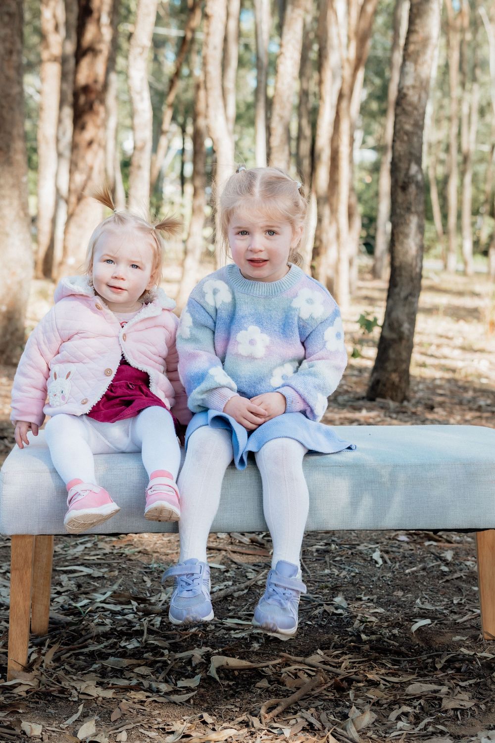 Two young blonde sisters sitting together on a bench outdoors in a forest, wearing pastel sweaters and dresses.