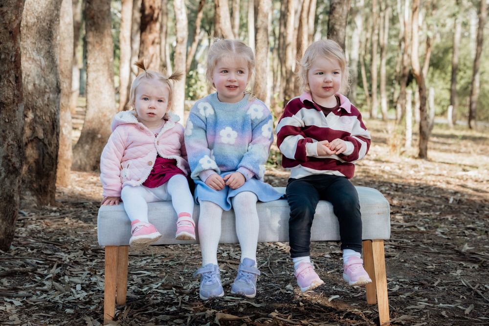 Three young children sitting together on a bench in a forest setting, wearing colorful outfits.