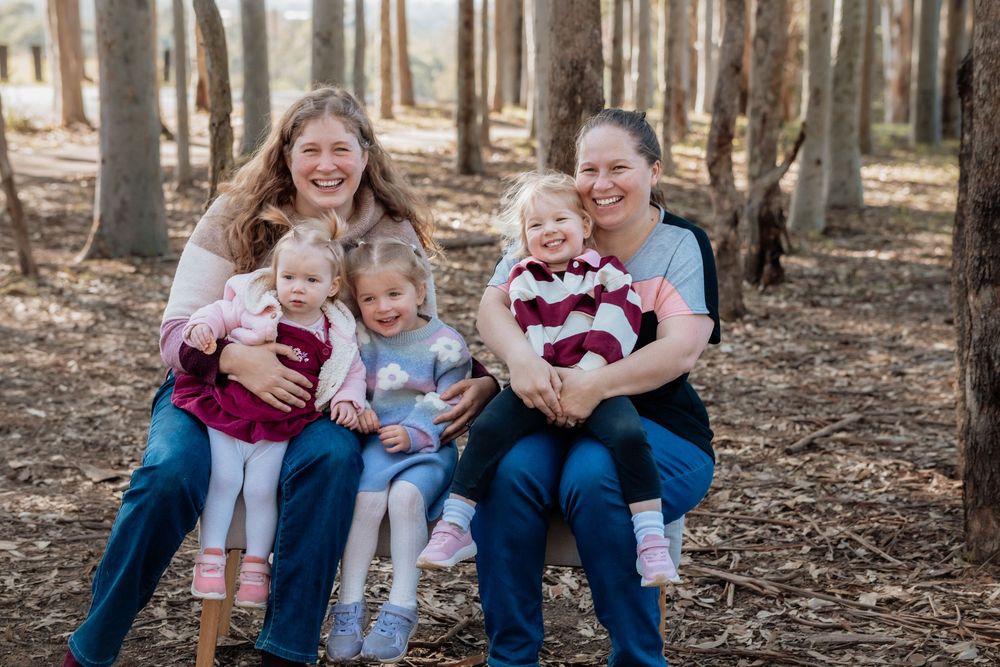 Two women sitting outdoors in a forest setting, smiling with three young girls on their laps during a family portrait.