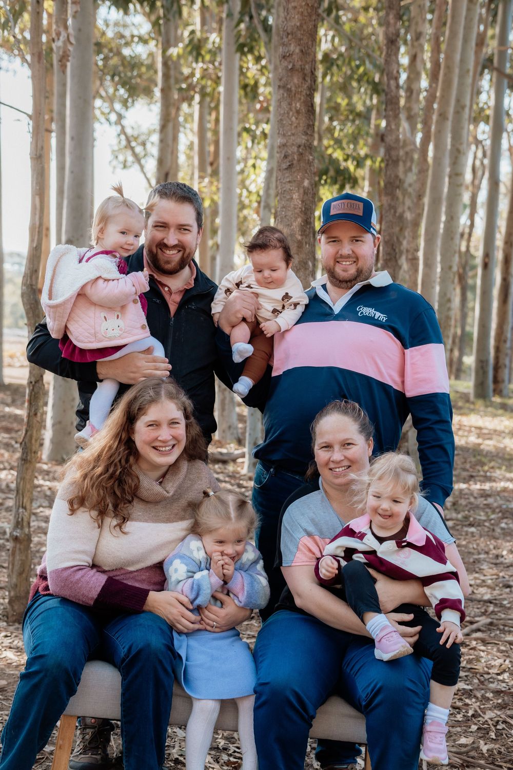 Two families with young children pose together outdoors among tall trees in a warm, candid group portrait.
