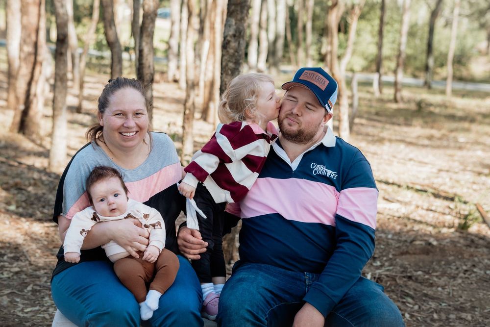 Family of four poses outdoors in wooded area, toddler kissing dad while mom holds baby at Rouse Hill Regional Park.