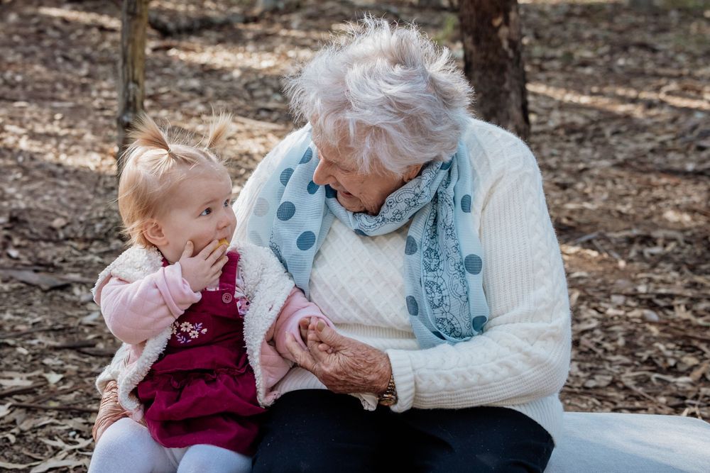 Elderly grandmother and toddler granddaughter sharing a tender moment together outdoors in a sunlit forest setting.