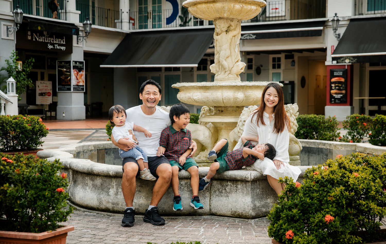 Family of four sitting and laughing together around a classic stone fountain outdoors.