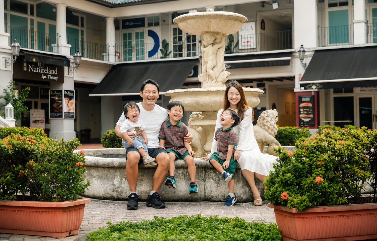Happy Asian family of five posing by a stone fountain in a charming outdoor shopping plaza.
