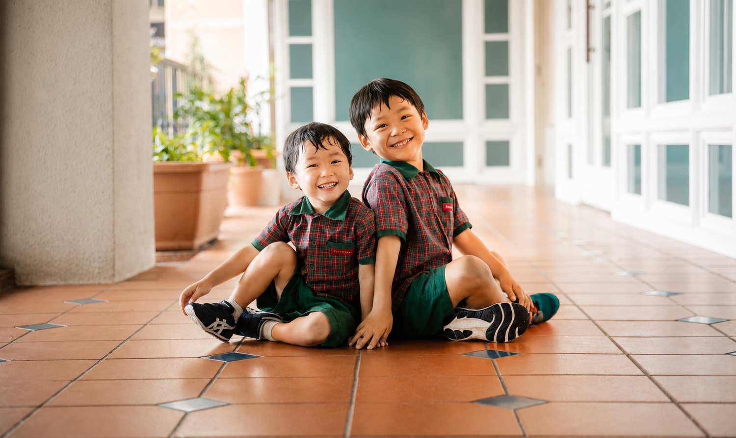 Two young Asian brothers in matching plaid shirts sitting back-to-back on terracotta tiles, smiling.