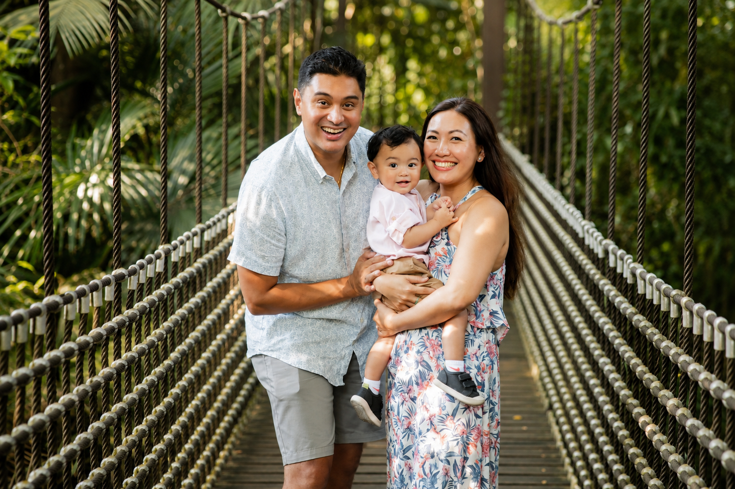 Smiling Asian couple holding their baby on a lush tropical suspension bridge in a garden.