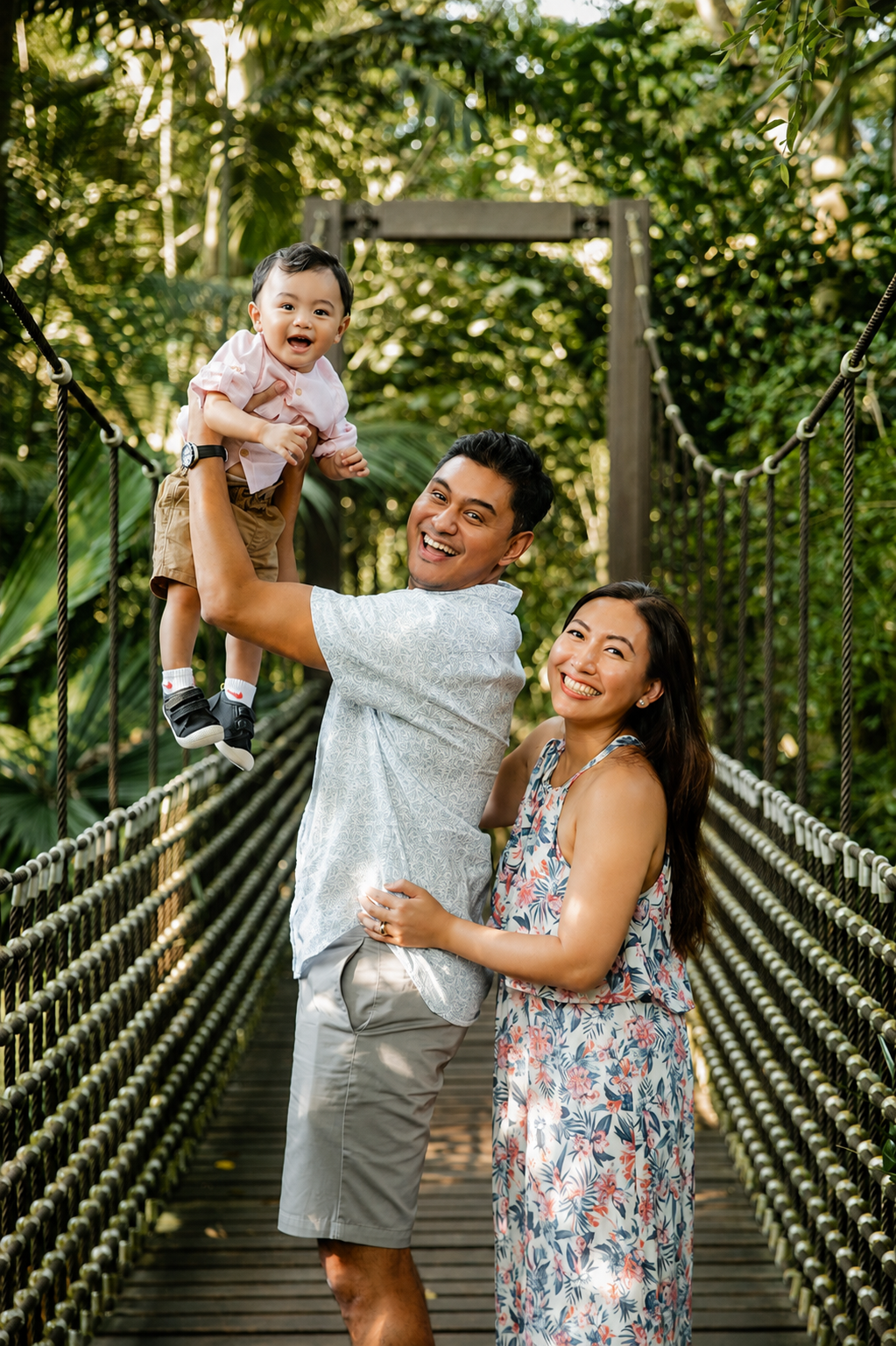 Joyful Asian father lifting laughing baby while mother smiles on a green tropical rope bridge.