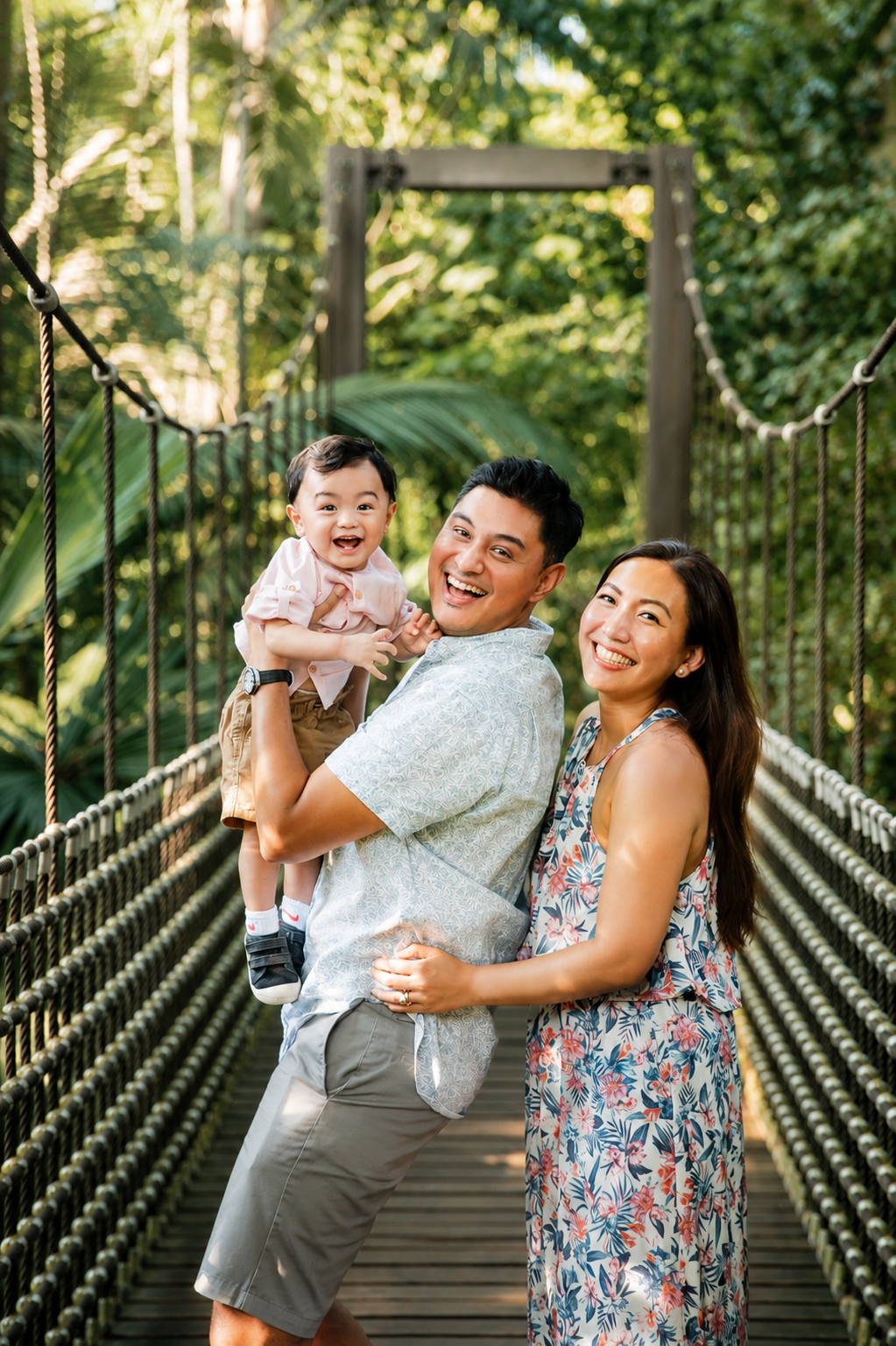 Happy Asian family of three posing together on a scenic rope suspension bridge in tropical greenery.