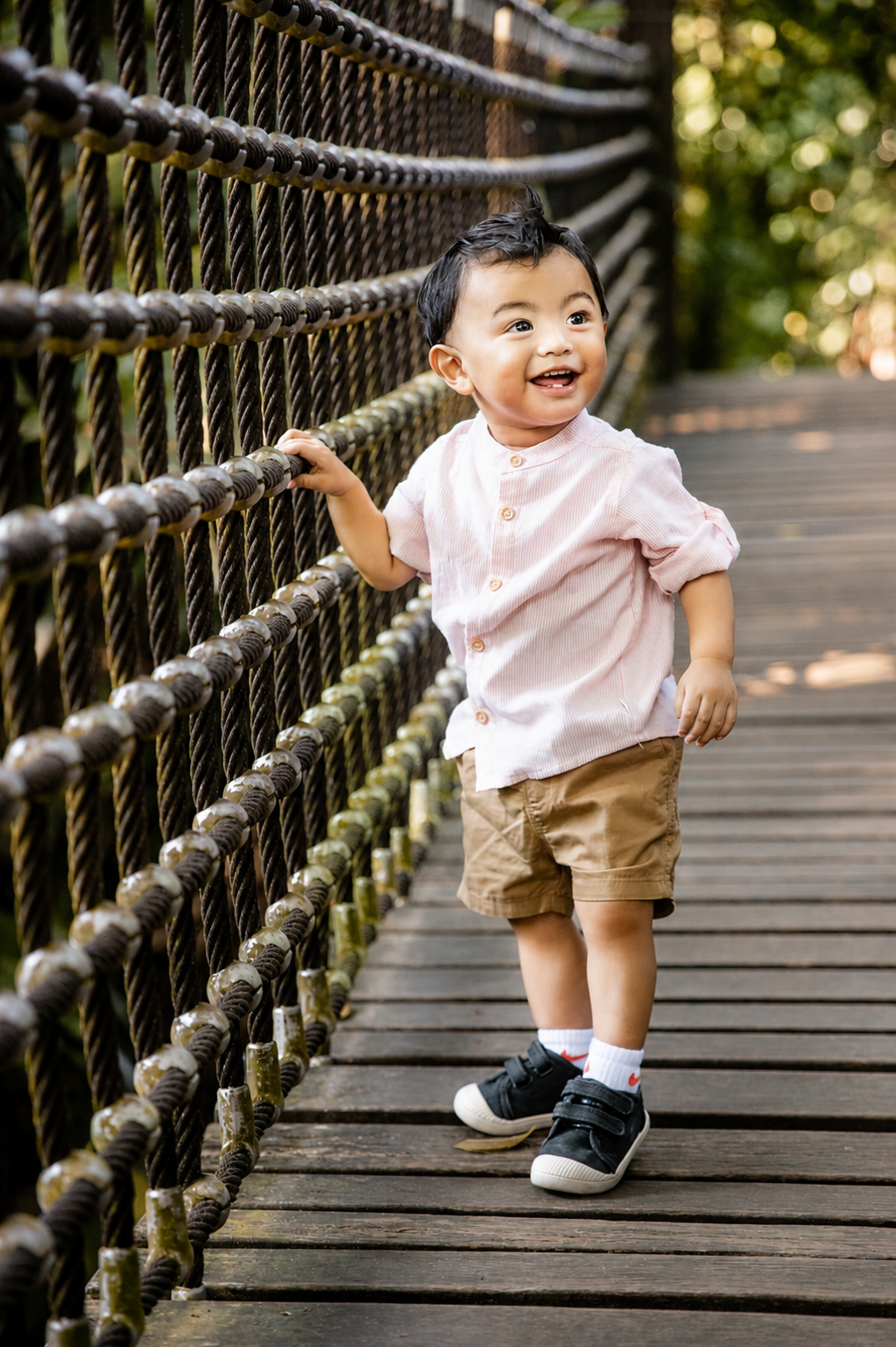 Adorable toddler in pink shirt and khaki shorts standing on a wooden rope bridge, laughing.
