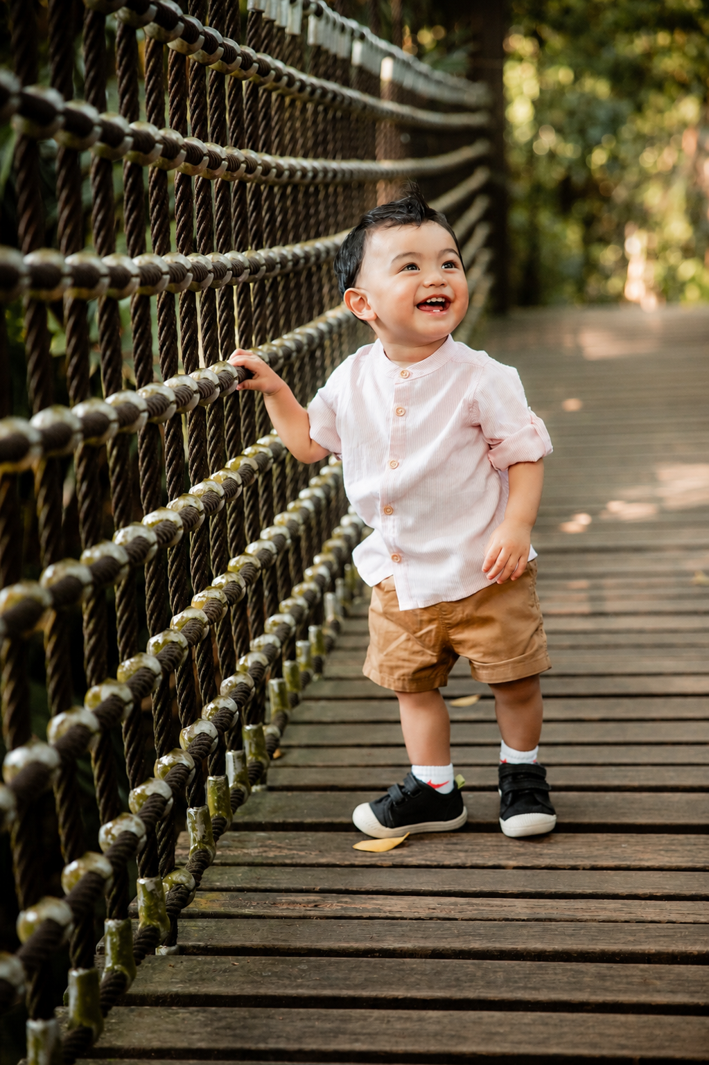 Cheerful baby boy in pink shirt holding rope railing on a wooden suspension bridge, smiling.