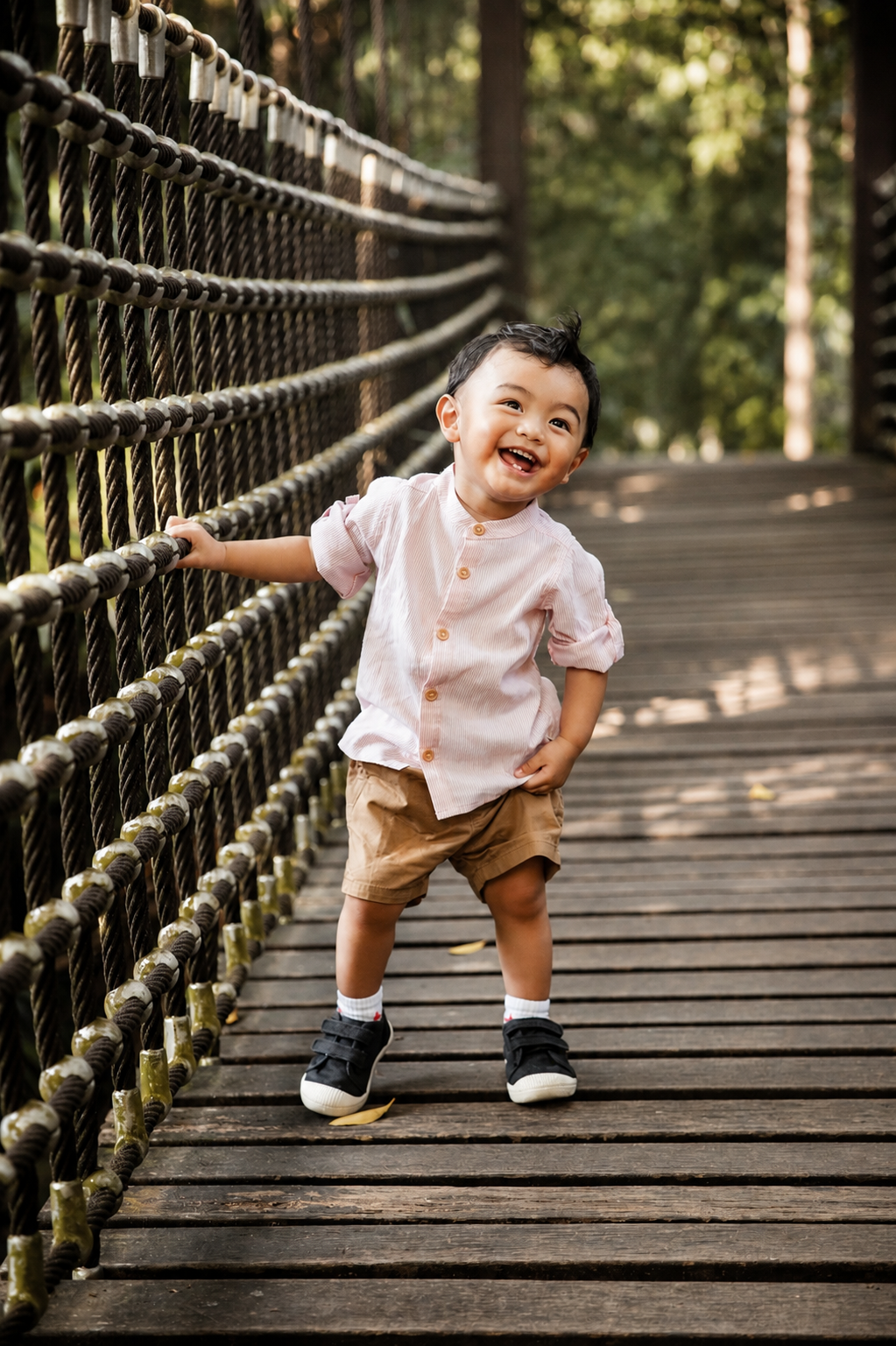 Happy toddler in a pink linen shirt and khaki shorts laughing on a wooden rope bridge.