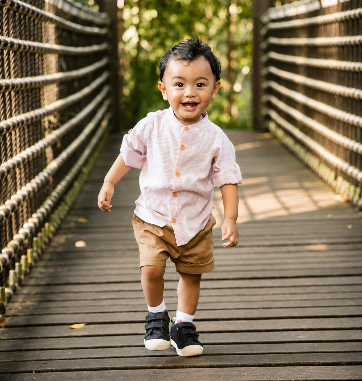 Cute Asian toddler in pink shirt and tan shorts walking confidently on a wooden suspension bridge.