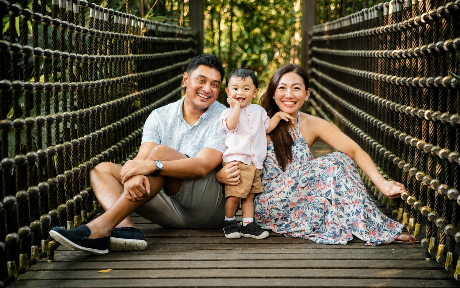 Happy Asian family sitting on wooden rope bridge, parents smiling with cheerful toddler between them in lush park.