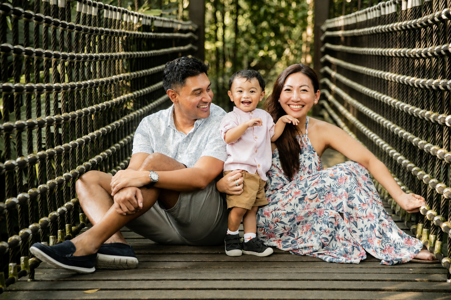 Joyful family of three sitting on wooden suspension bridge, parents laughing with excited baby in outdoor setting.