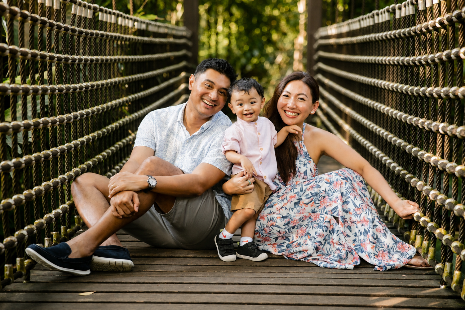 Loving Asian family posing on rope bridge in forest, mom and dad holding smiling toddler in natural setting.