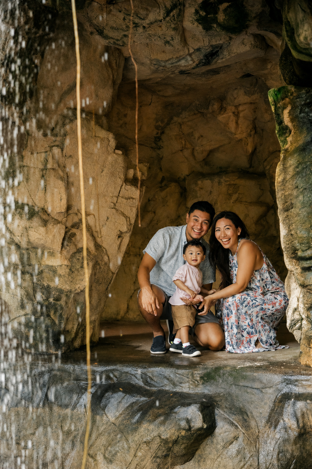 Family of three posing beside waterfall in rocky cave, parents crouching with baby in scenic natural grotto.