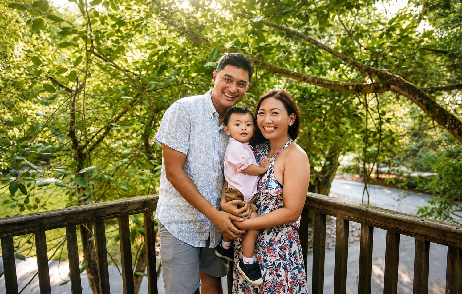 Young Asian family standing on wooden deck holding toddler, surrounded by lush green trees in park setting.
