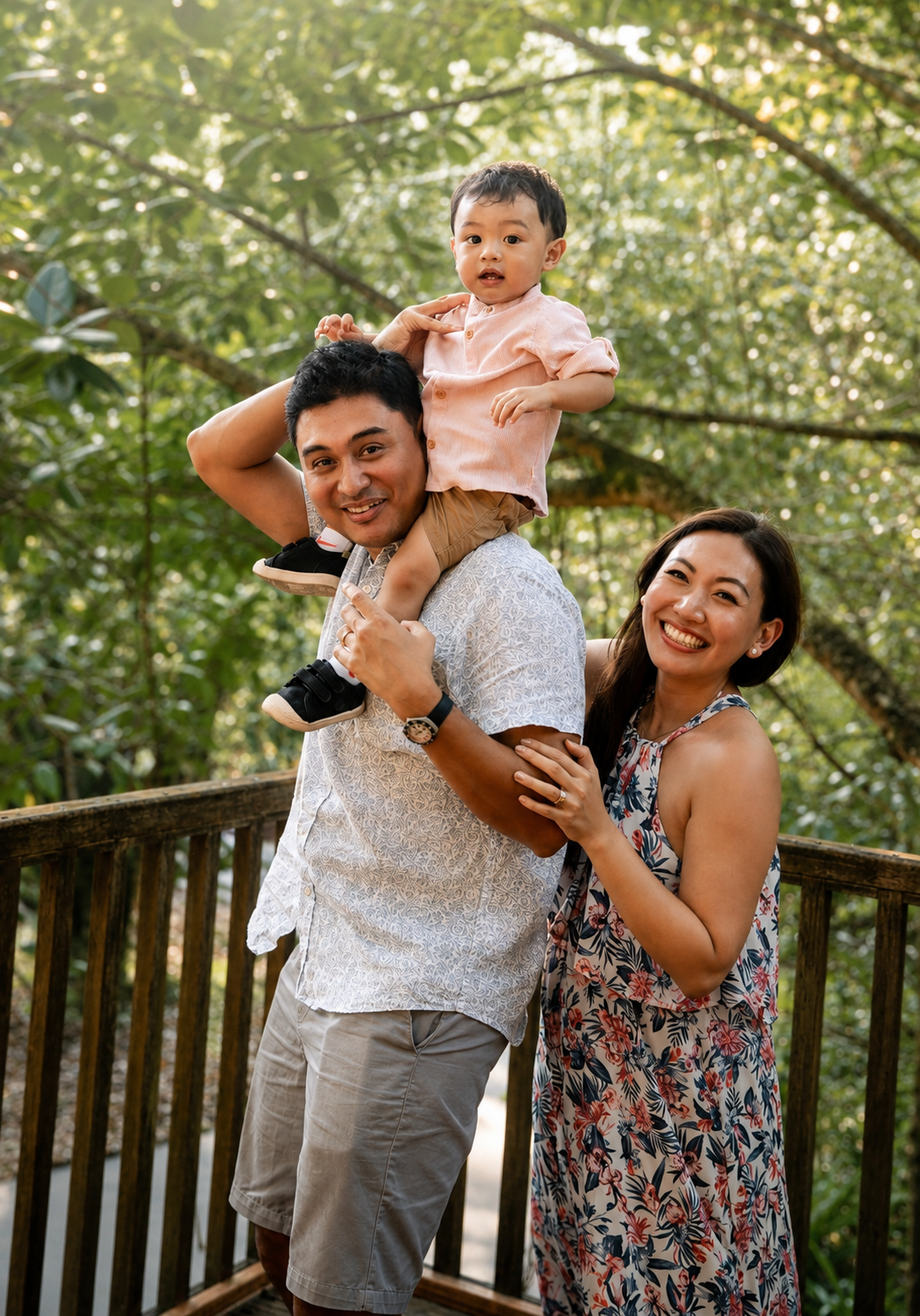 Father carrying toddler on shoulders with mother beside them, all smiling on wooden bridge in green forest.