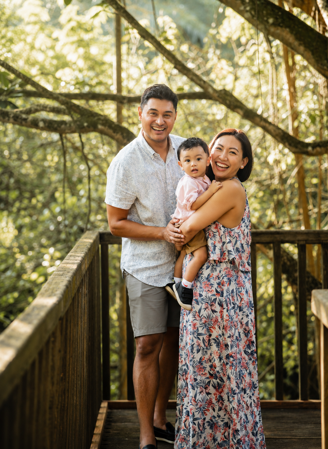 Smiling Asian family posing on wooden bridge, father and mother holding baby girl in sunlit forest setting.