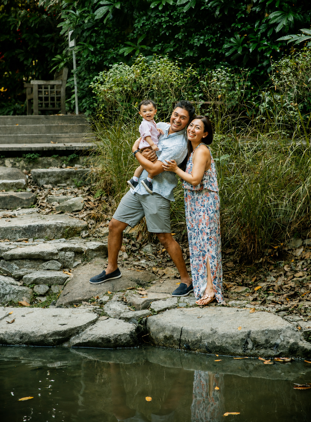 Happy family of three posing on stepping stones by pond, father holding baby with mother in floral dress.