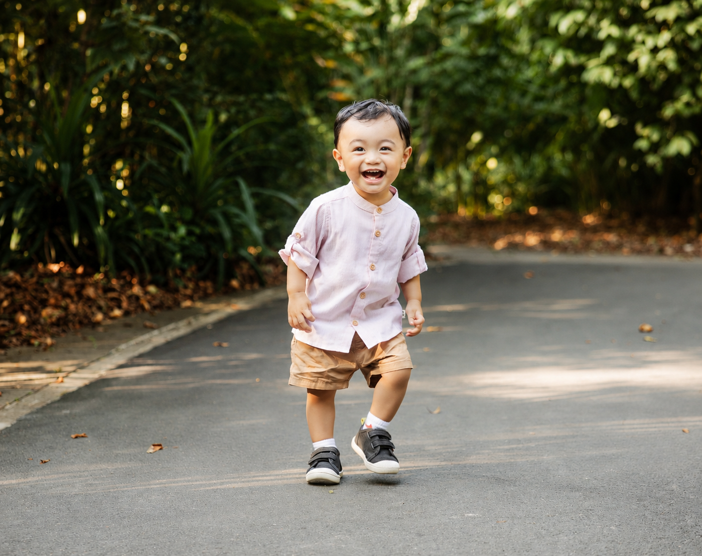 Adorable toddler boy in pink shirt and khaki shorts laughing while running on park path with greenery behind.