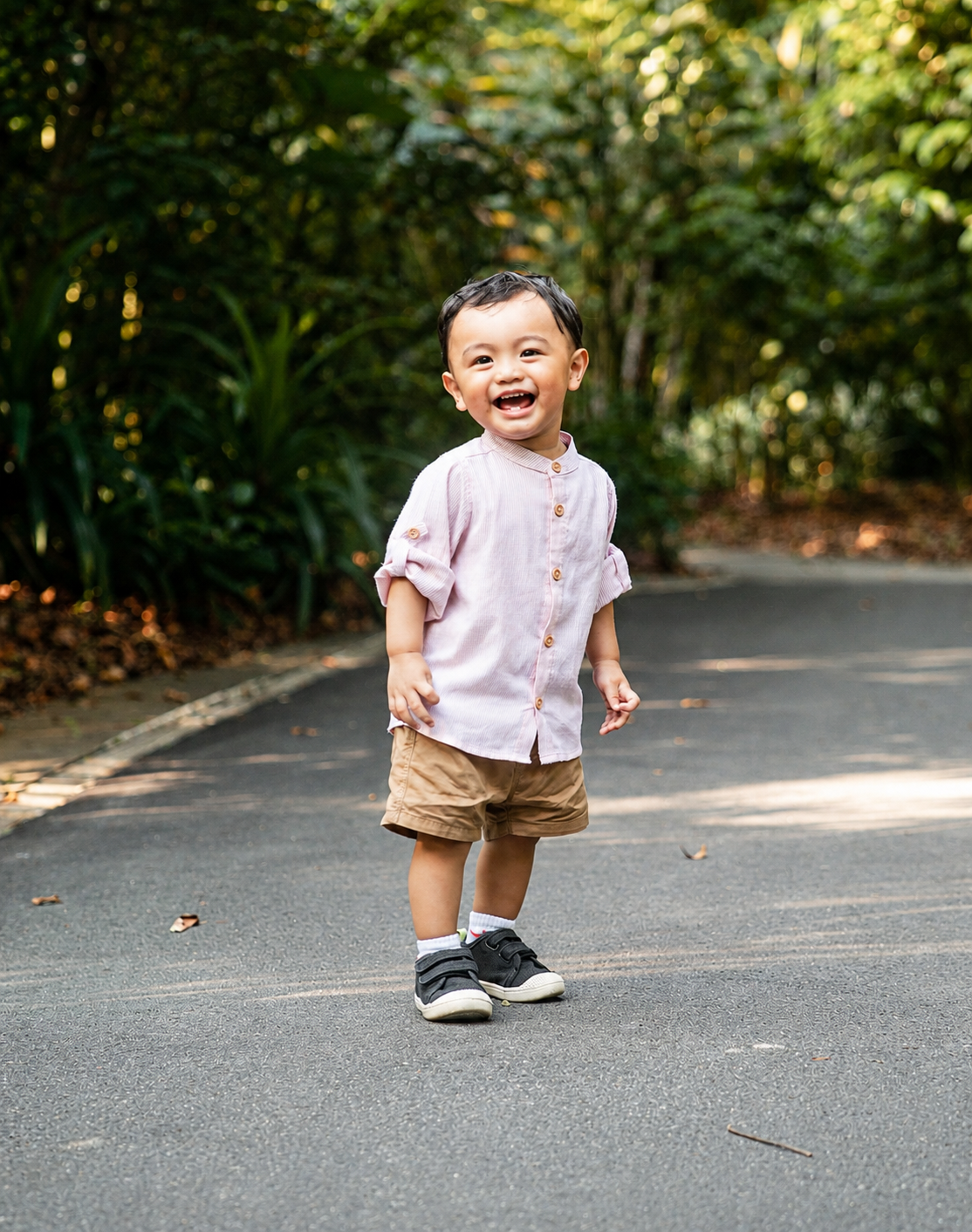Cute Asian toddler boy in light pink shirt and tan shorts smiling while walking on sunny park pathway.