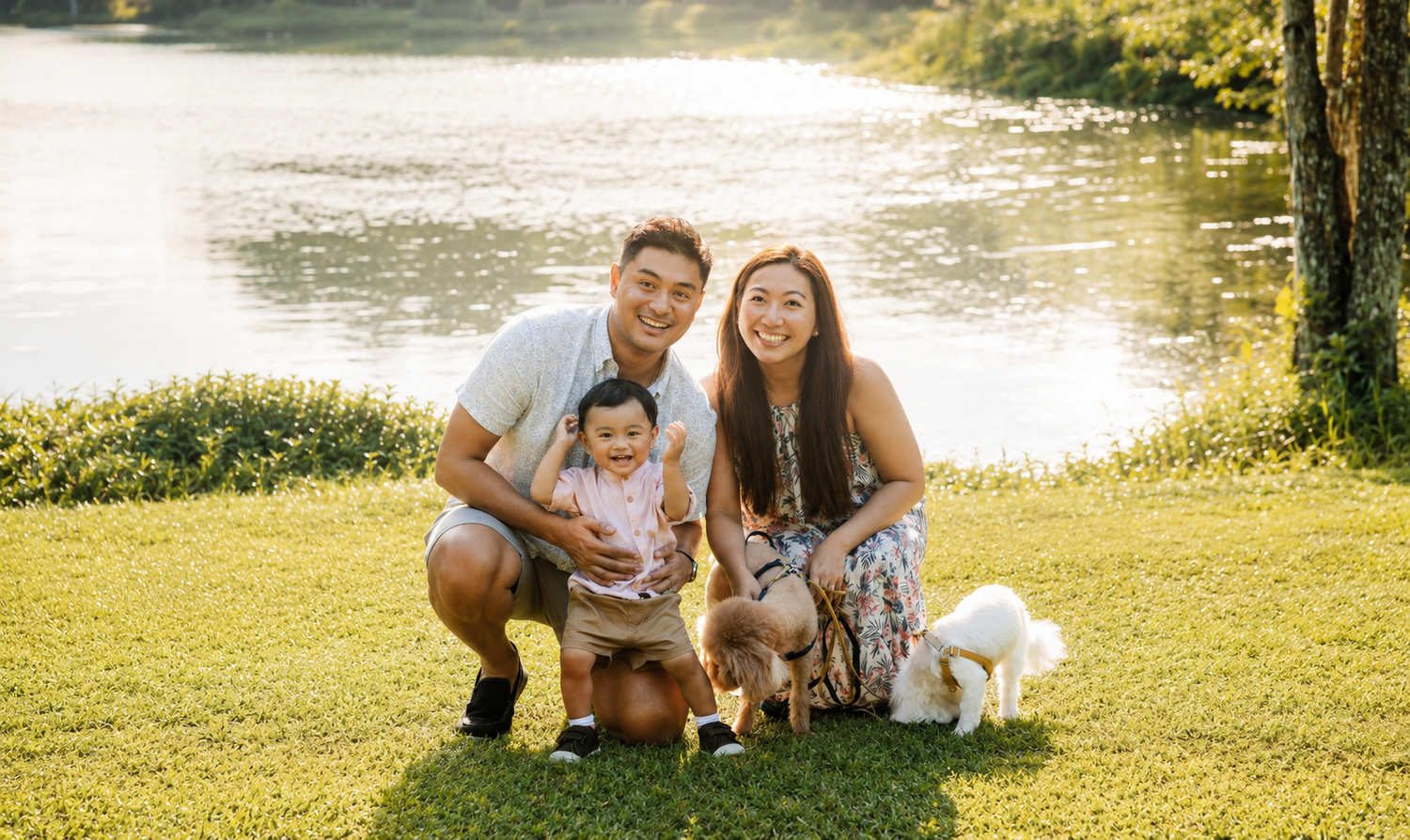 Family with baby and white dog posing by a sunlit lake on green grass.
