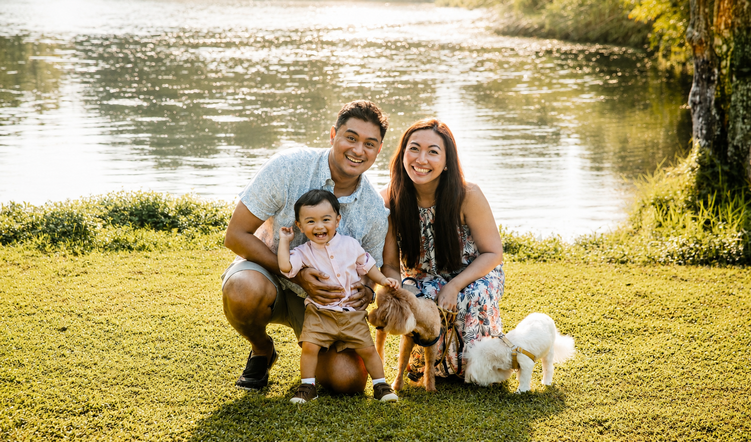 Smiling family with infant and two dogs sitting on grass by a peaceful lake.