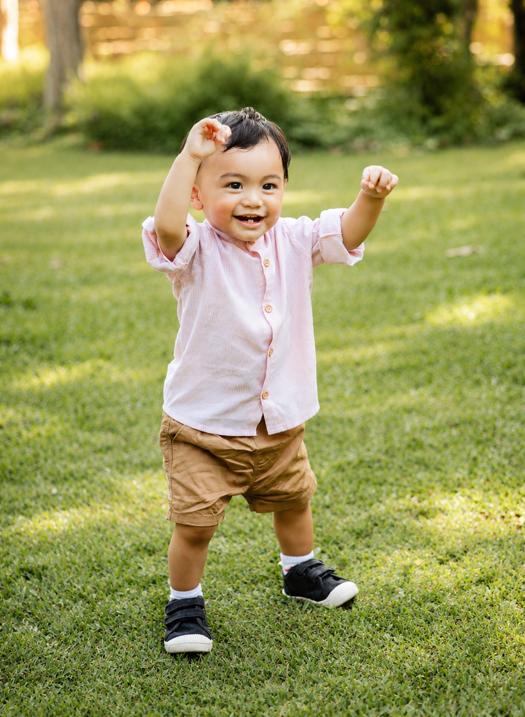 Cheerful baby boy in pink shirt raising arms while standing on green lawn.