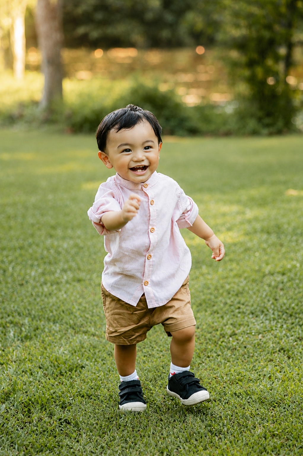 Smiling toddler in pink button shirt and khaki shorts walking on green grass.