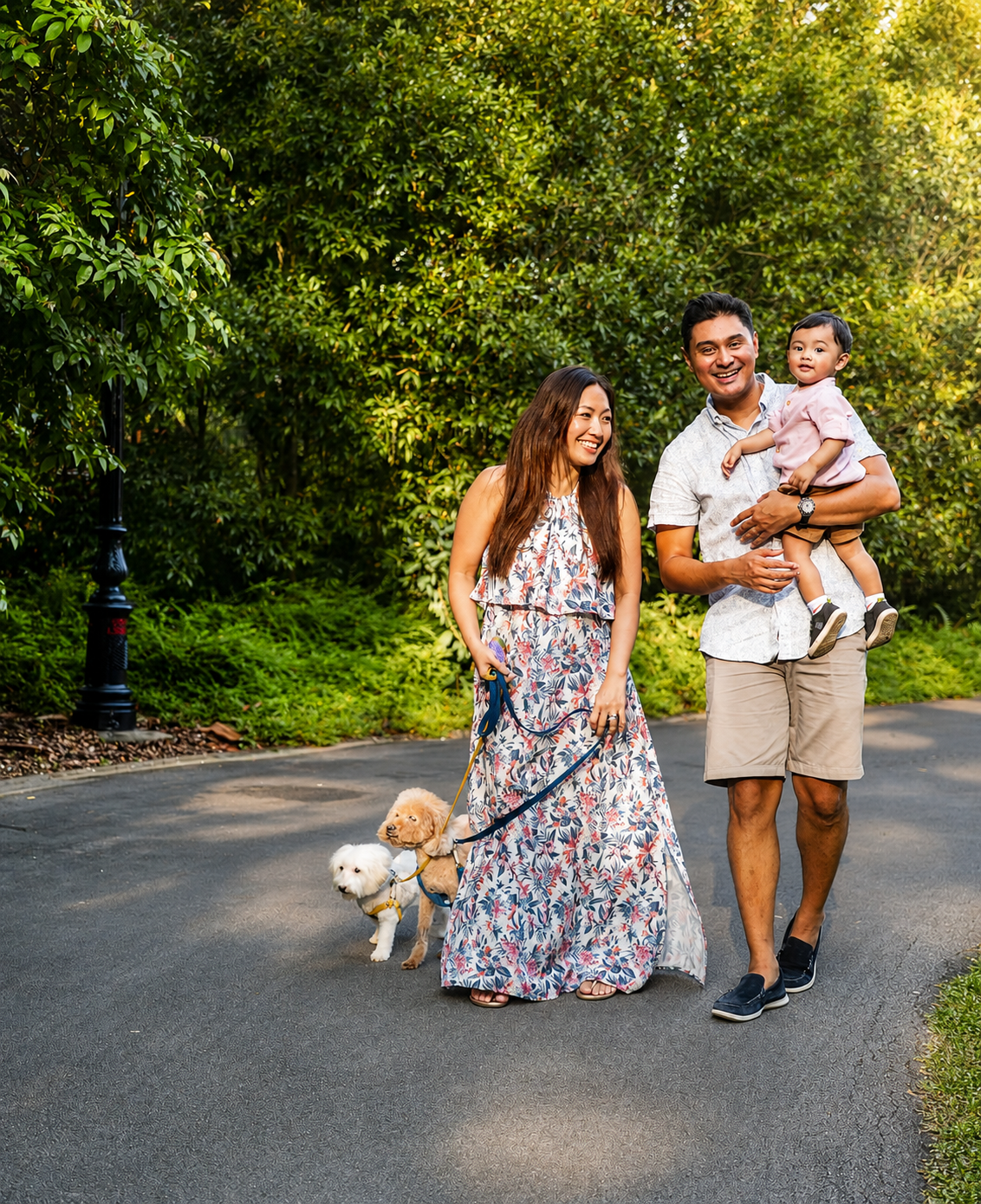Family with baby and dog on leash walking along a tree-lined park path.