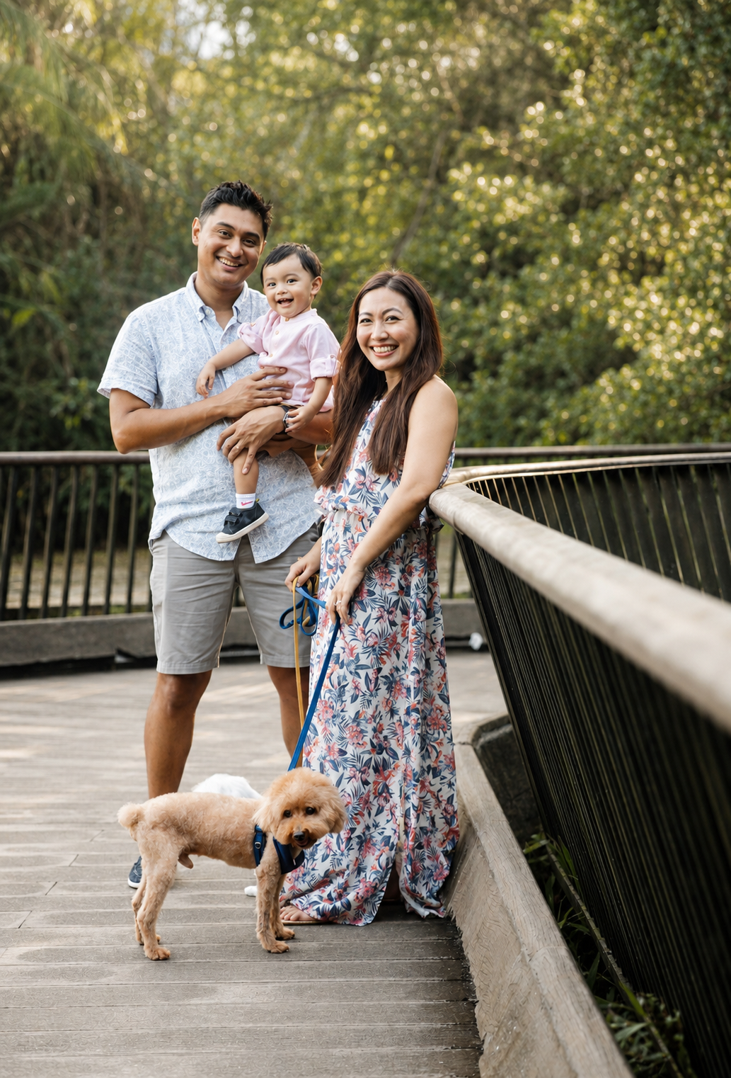 Smiling family holding baby and dog on leash posing on a wooden bridge.