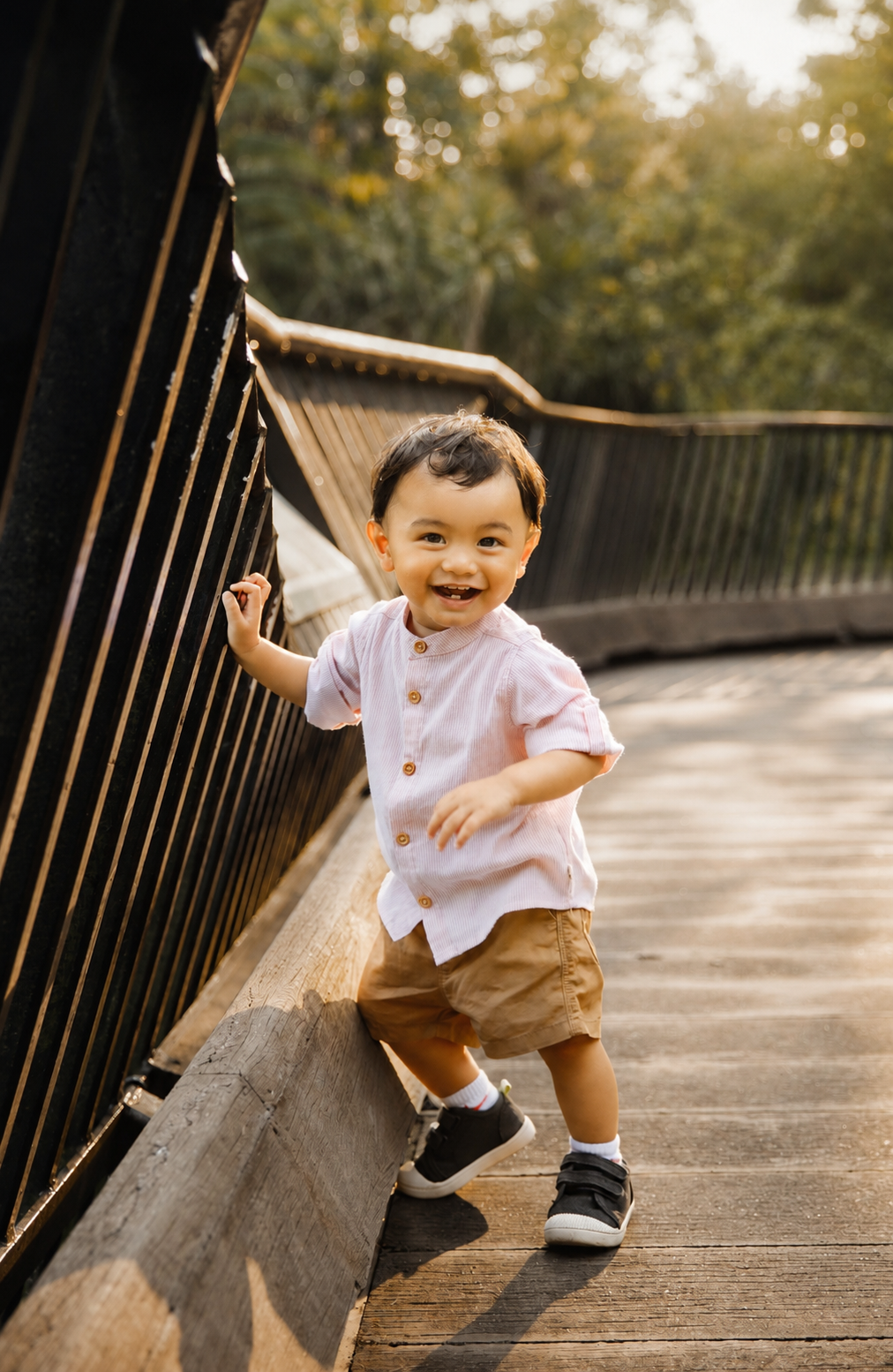 Happy toddler boy in pink shirt and khaki shorts laughing while walking on a sunny wooden bridge.