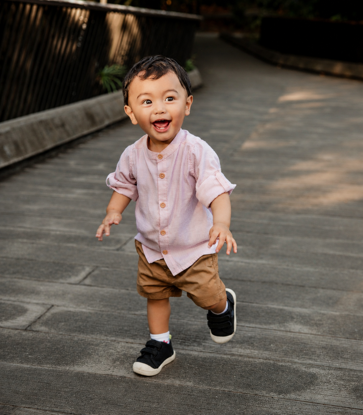Joyful toddler boy in lavender shirt and tan shorts running and laughing on an outdoor pathway.