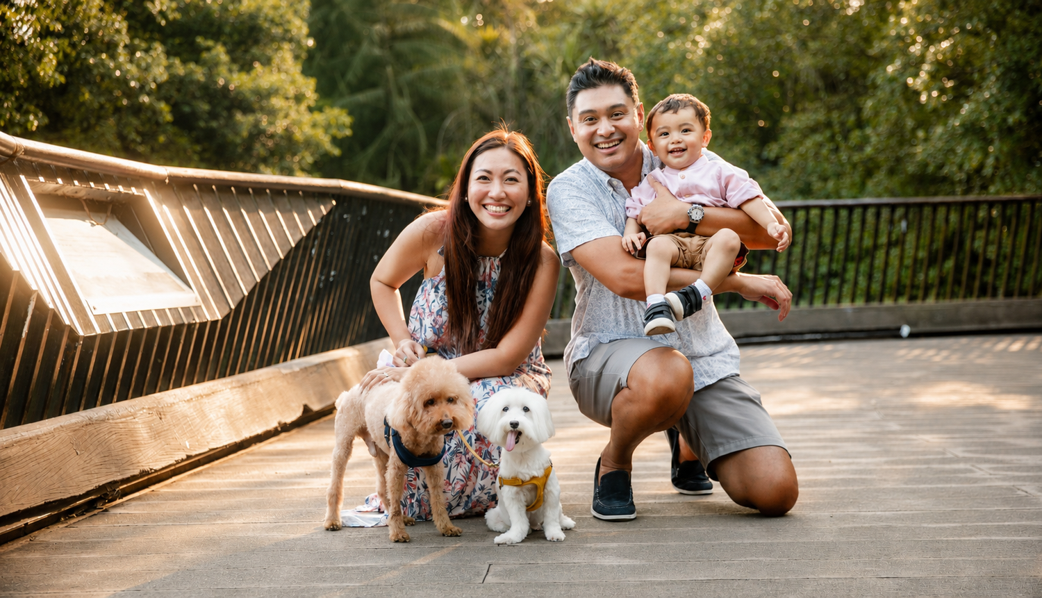 Smiling Asian family with toddler boy and two small fluffy dogs posing on a wooden bridge outdoors.