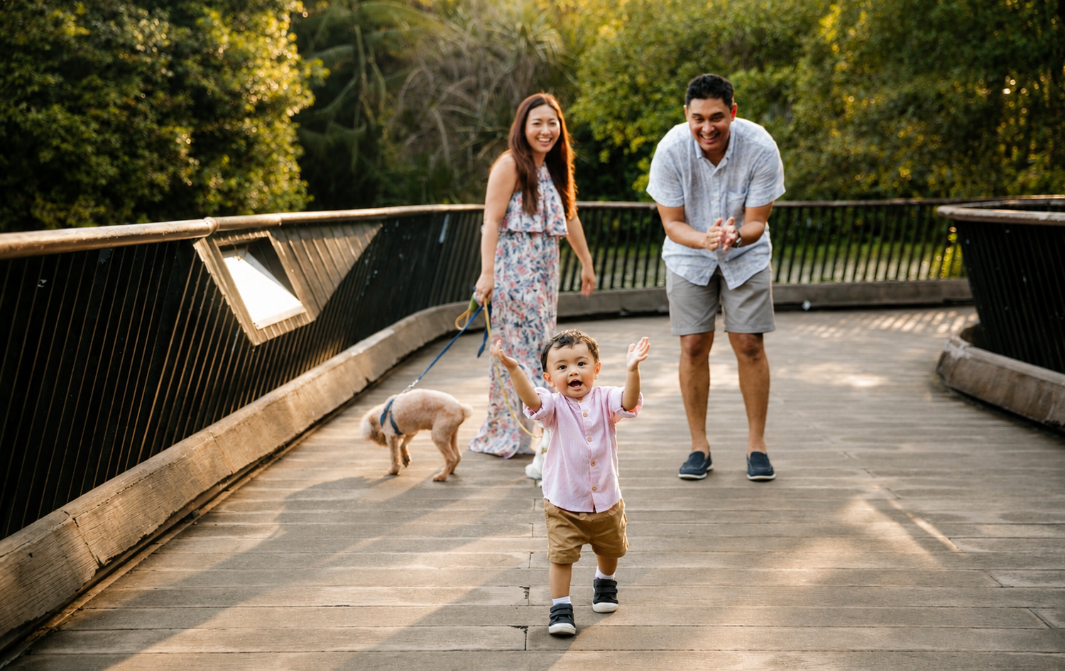 Parents laughing as their excited toddler boy runs ahead on a sunny wooden bridge with a dog.