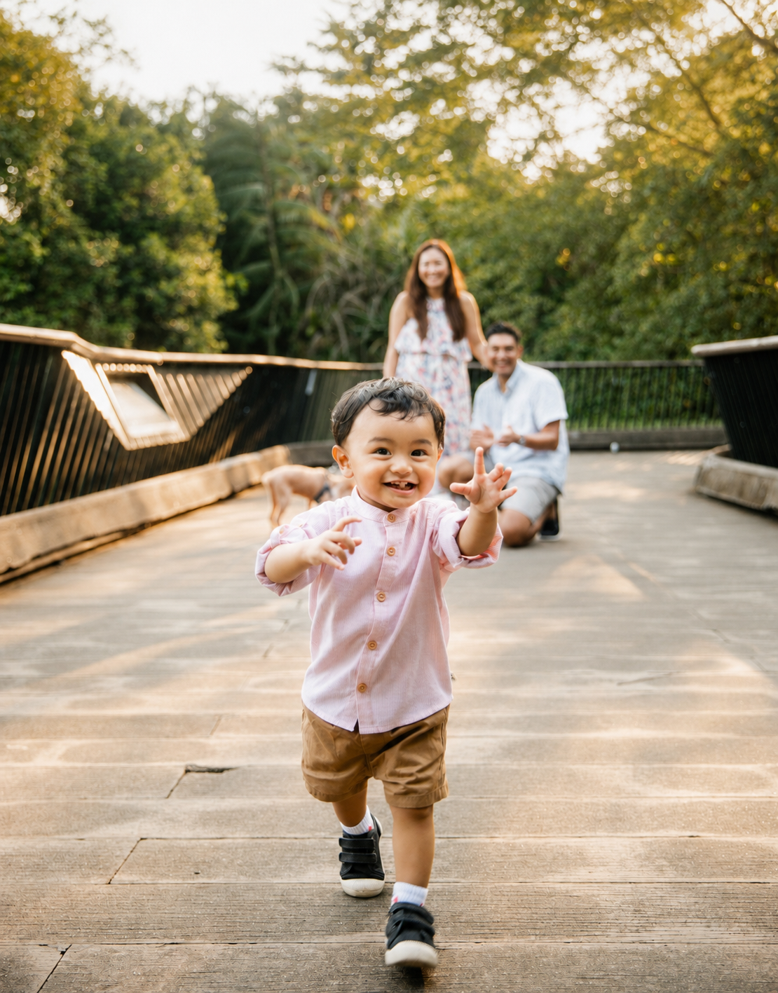 Smiling toddler boy running toward camera with arms out as parents kneel on a sunny wooden bridge.