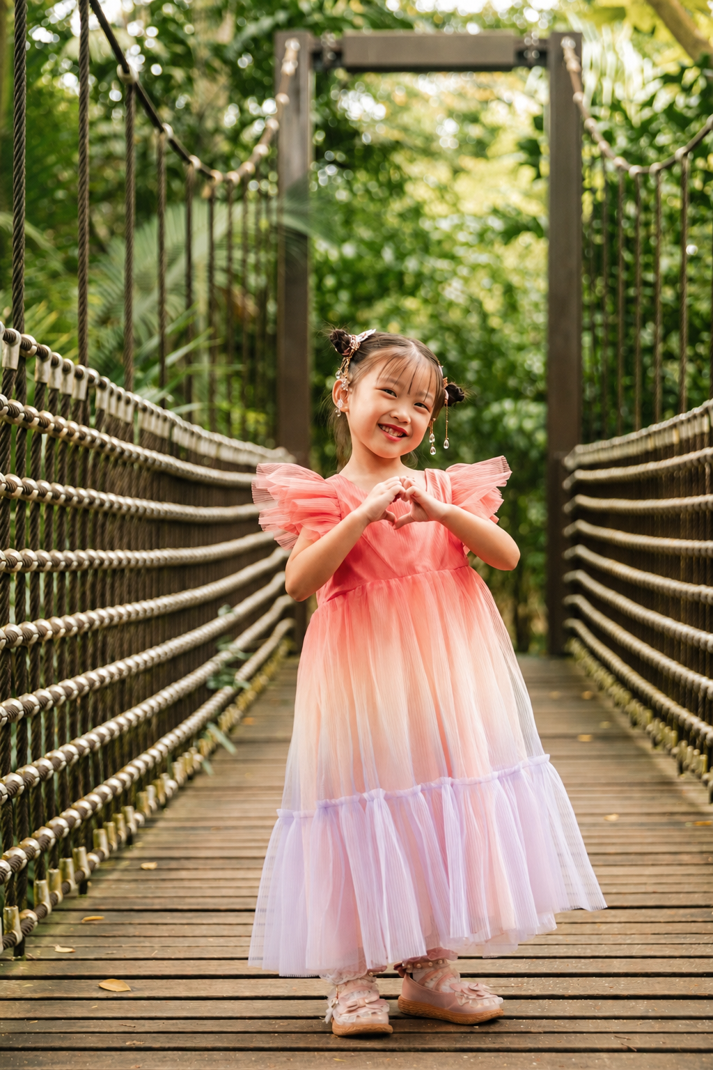 Adorable little girl in a pink gradient tulle dress making a heart shape with her hands on a rope bridge.