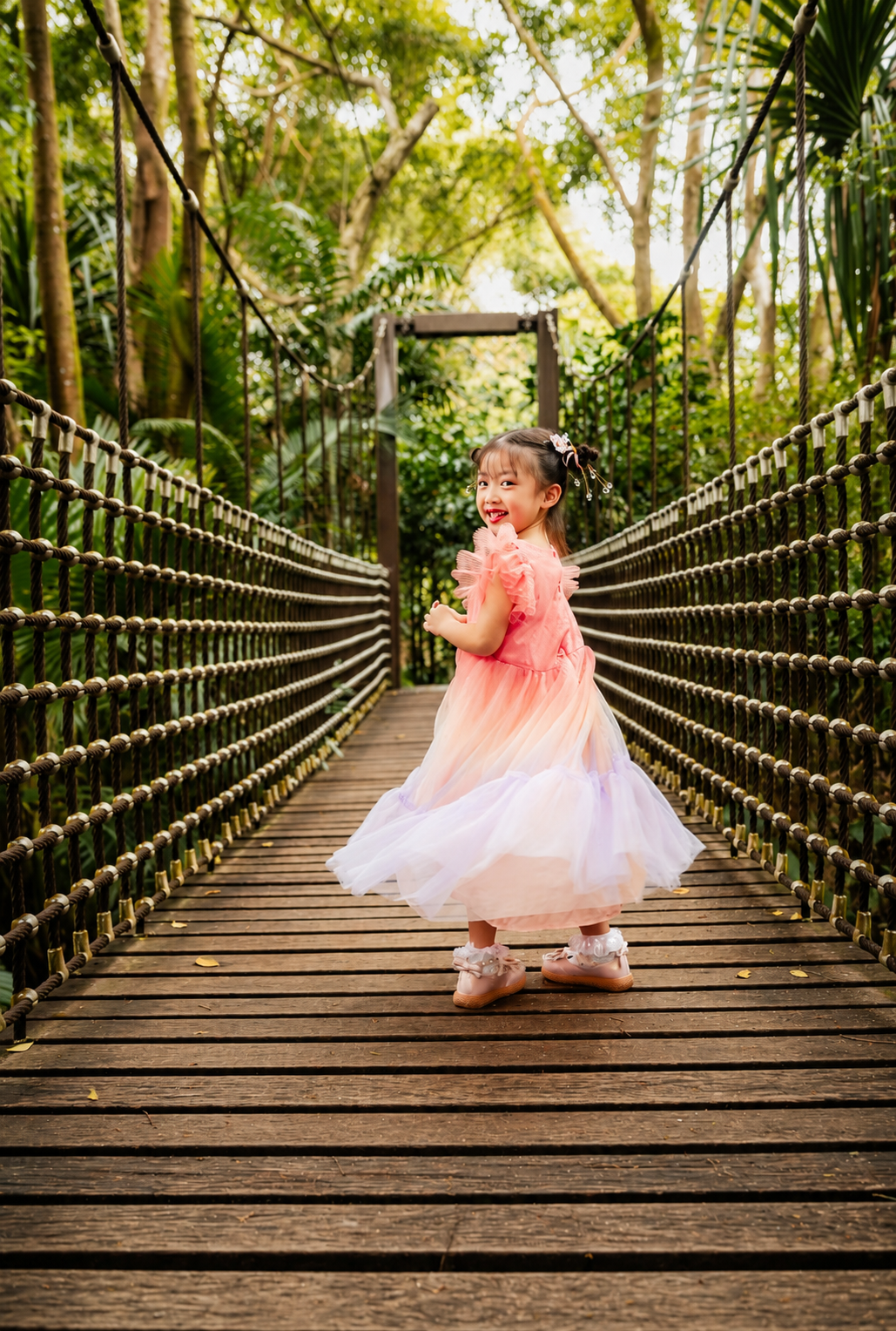 Young girl in pink tulle dress twirling on a wooden suspension bridge surrounded by lush greenery.