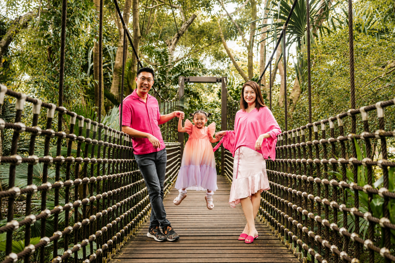 Happy family in matching pink outfits playing on a wooden suspension bridge in a tropical forest.