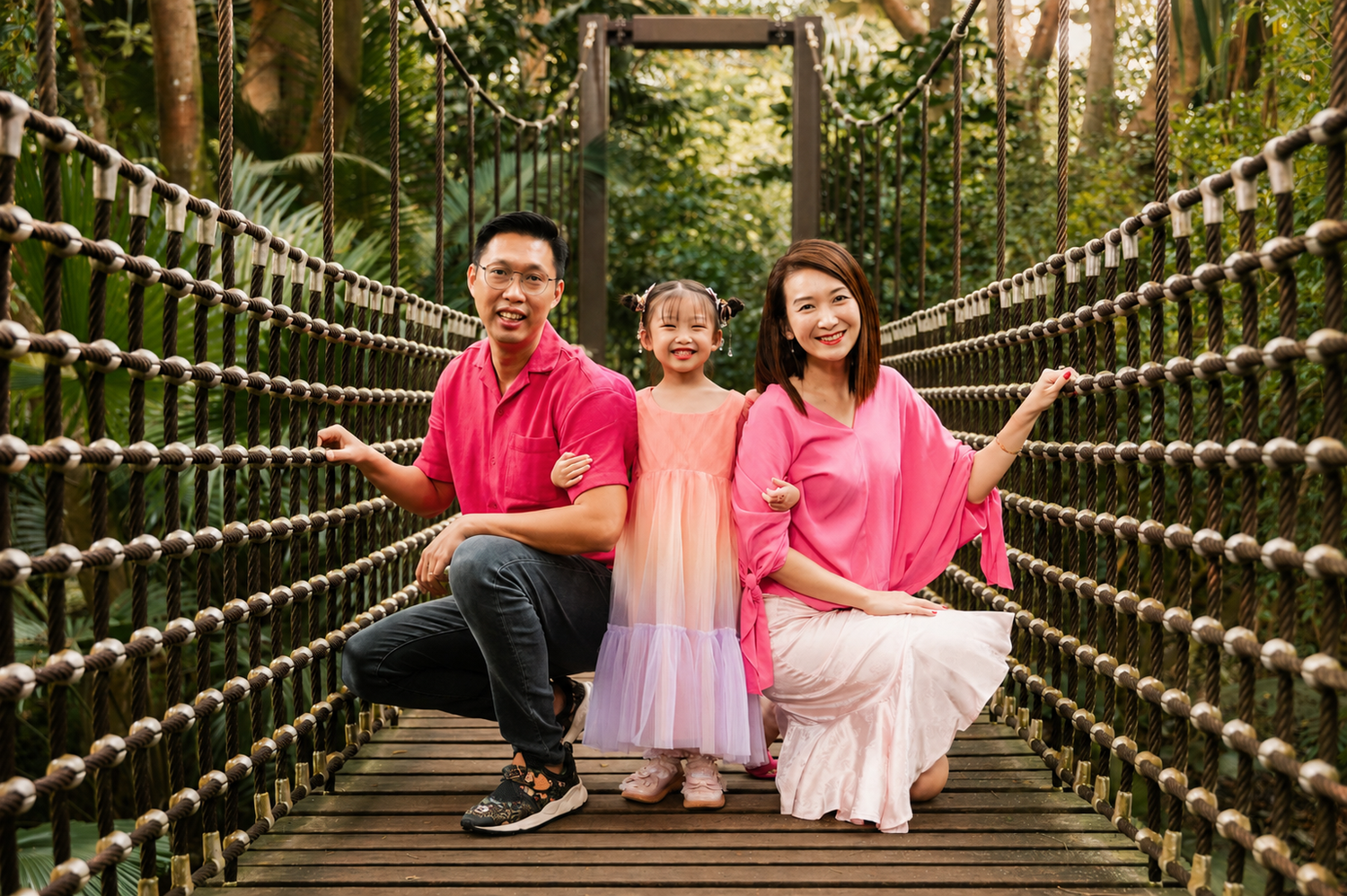 Smiling family of three posing together on a moss-covered wooden suspension bridge in a green park.