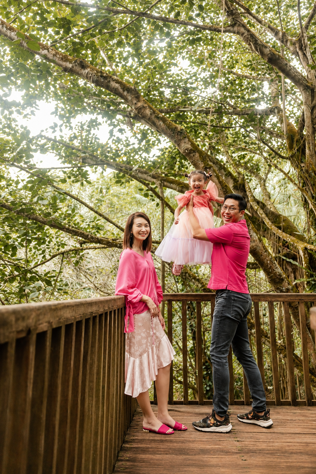 Father lifting daughter in pink dress while mother watches on a wooden deck surrounded by trees.