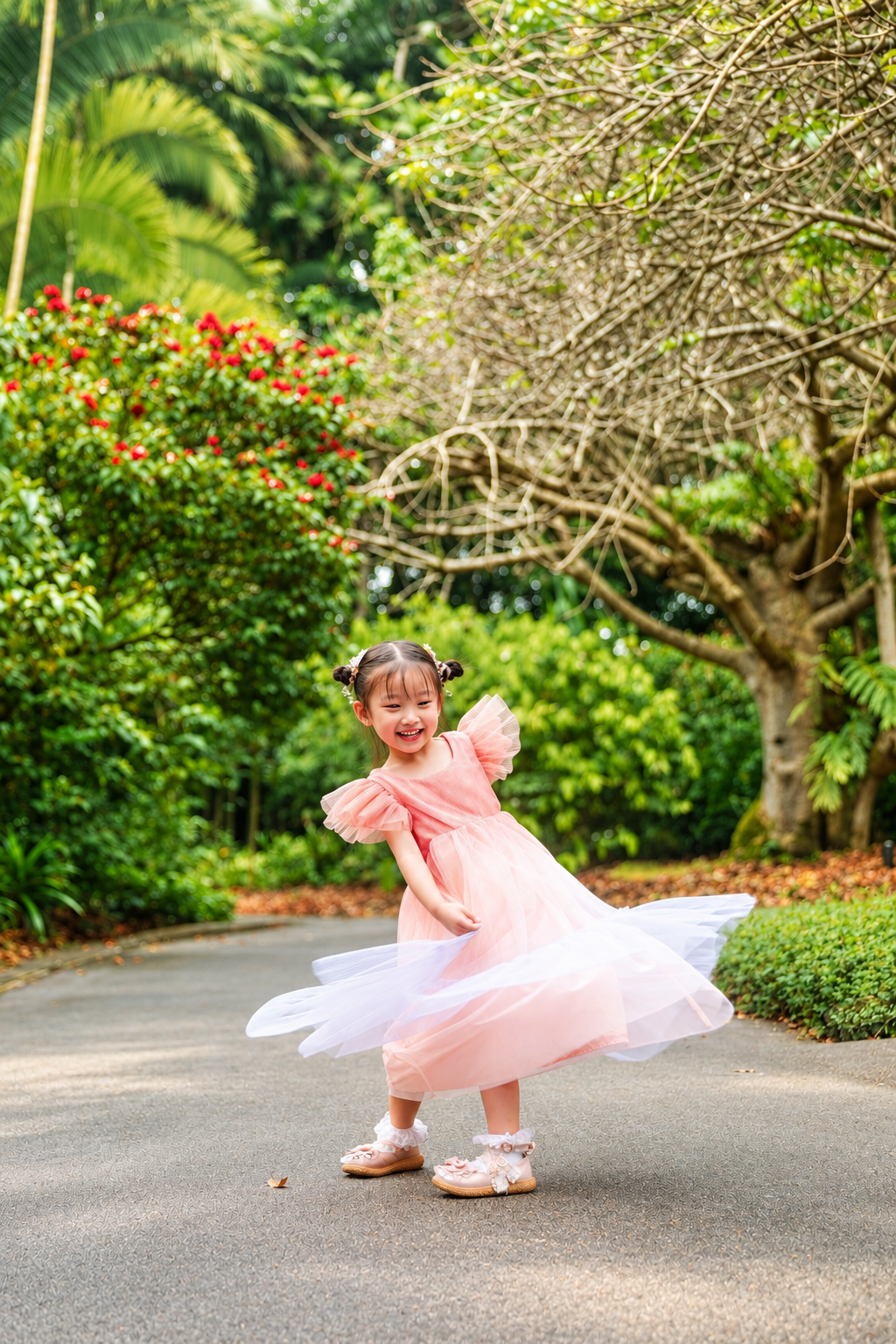 Joyful young girl in pink tulle dress spinning on a tree-lined park pathway with colorful flowers.